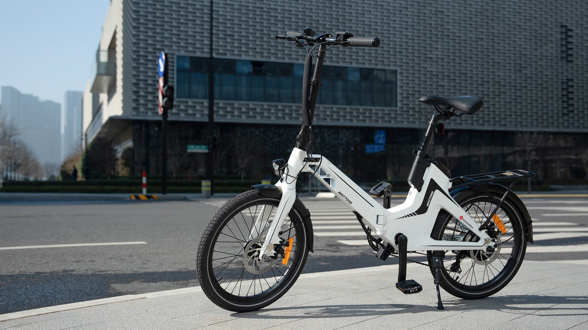 A white electric bicycle stands on a city sidewalk, with a modern building and traffic lights