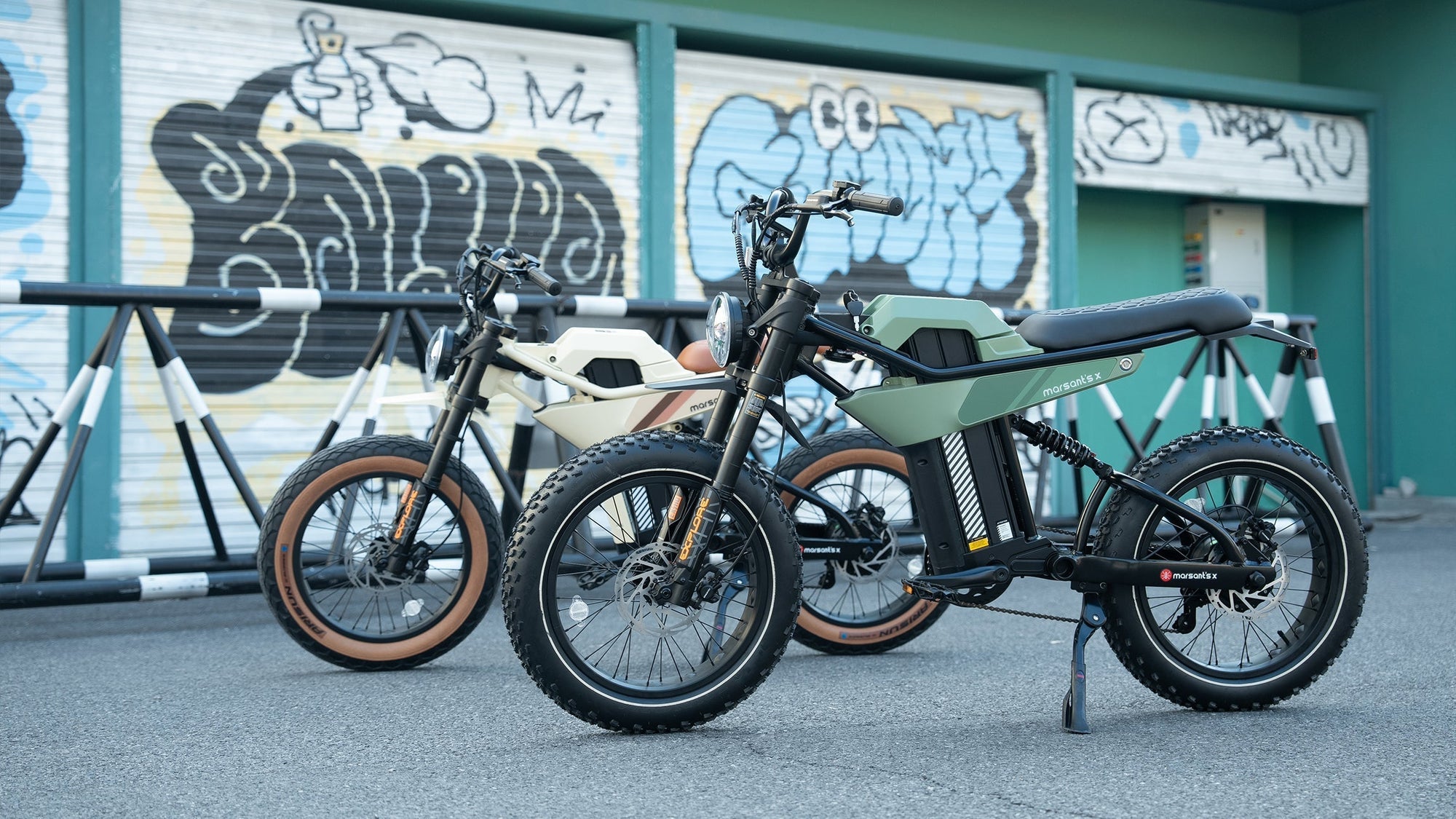 Two Marsant's X electric bikes with fat tires are parked in front of a colorful graffiti wall.