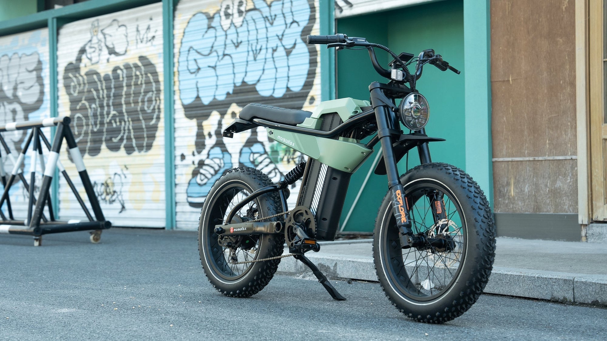 A sleek electric bike with thick tires is parked on a street, set against a backdrop of colorful graffiti.