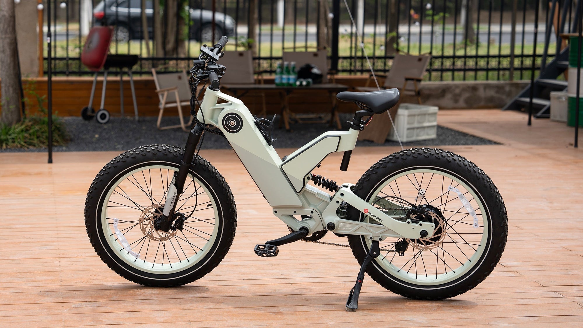 A sleek white electric bike with thick tires is parked on a tiled patio, near outdoor furniture and greenery.