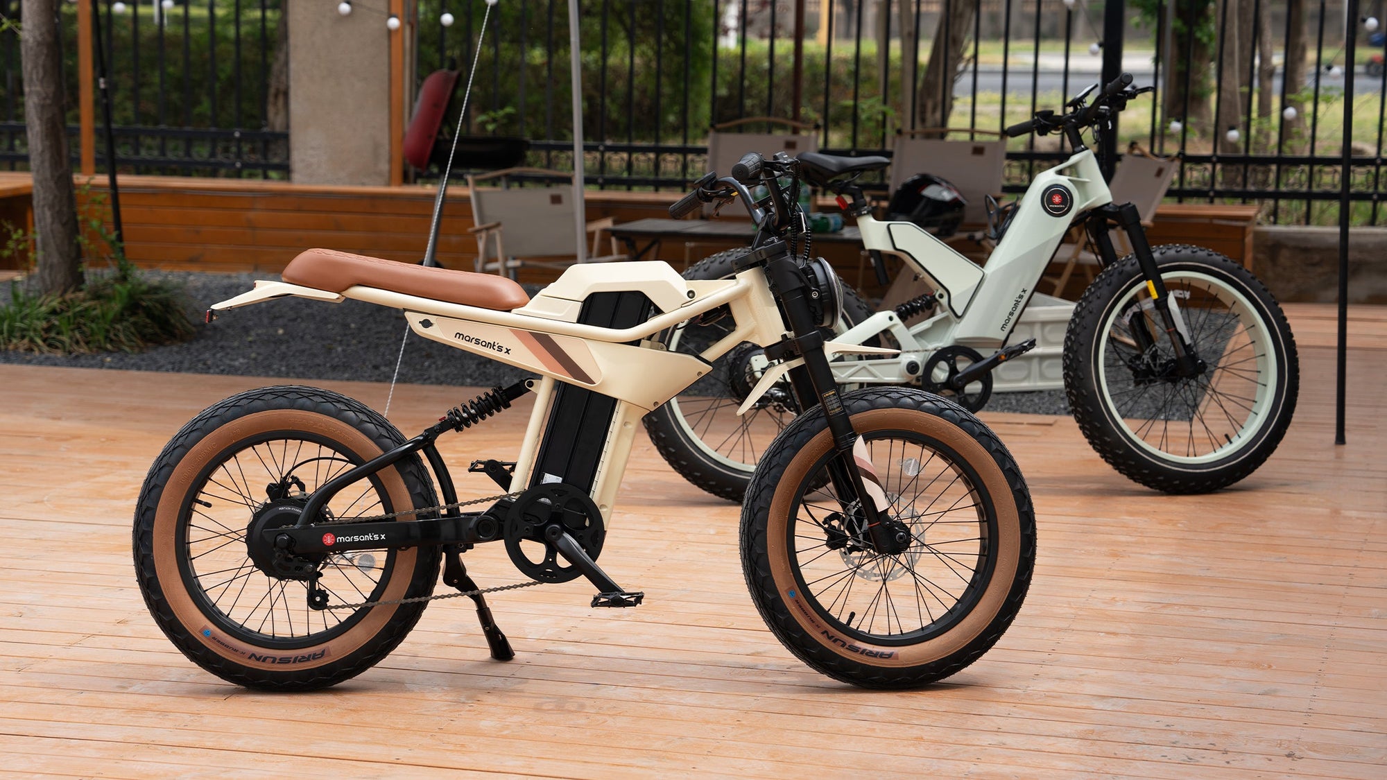Two stylish electric bikes with thick tires and modern designs are parked on a wooden deck in an outdoor setting.