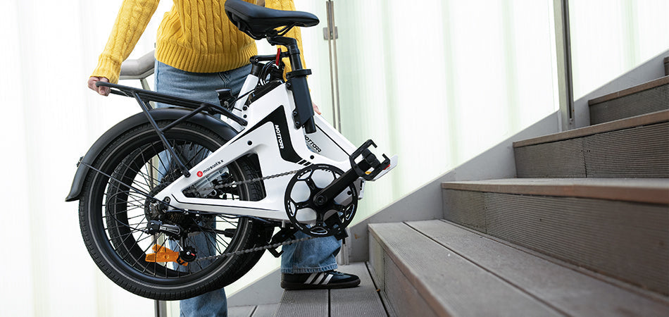  A person in a yellow sweater carries a white folding bike up wooden stairs, showcasing its compact design.