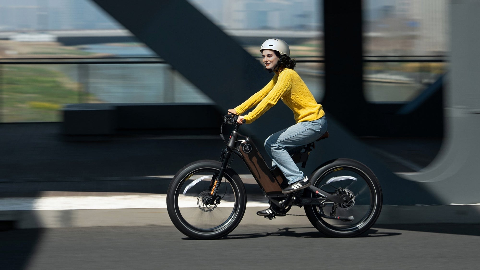 A woman in a yellow sweater and helmet rides an electric bike, smiling as she moves through a modern urban setting.
