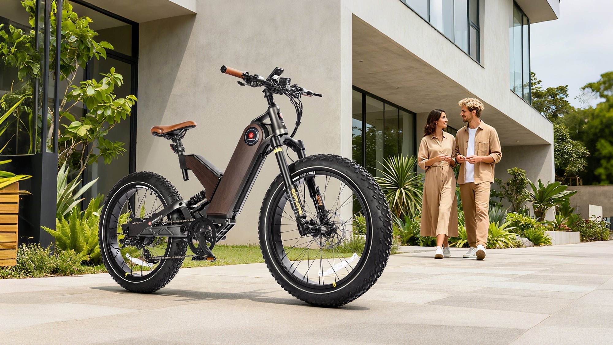 A modern electric bike with fat tires stands in front of a stylish house, as a couple walks by in matching outfits.