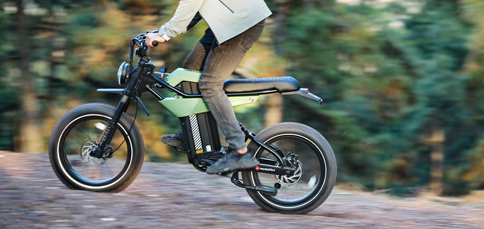 A person rides a green electric bike on a forest path, wearing jeans and a beige jacket.